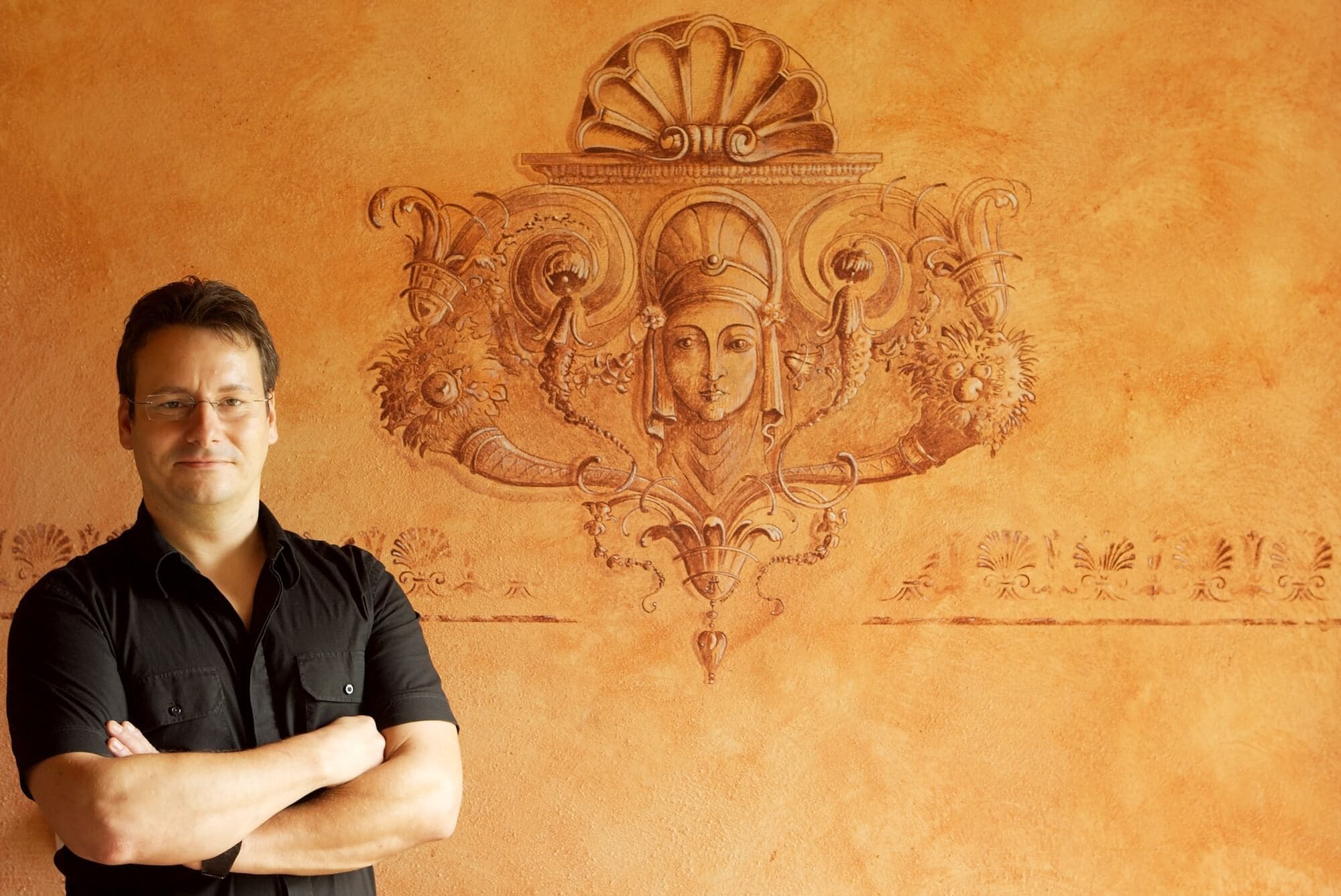 Toni Hamler standing in front of an intricate wall painting featuring classical ornamental motifs and a female figure, showcasing his expertise in decorative wall techniques.